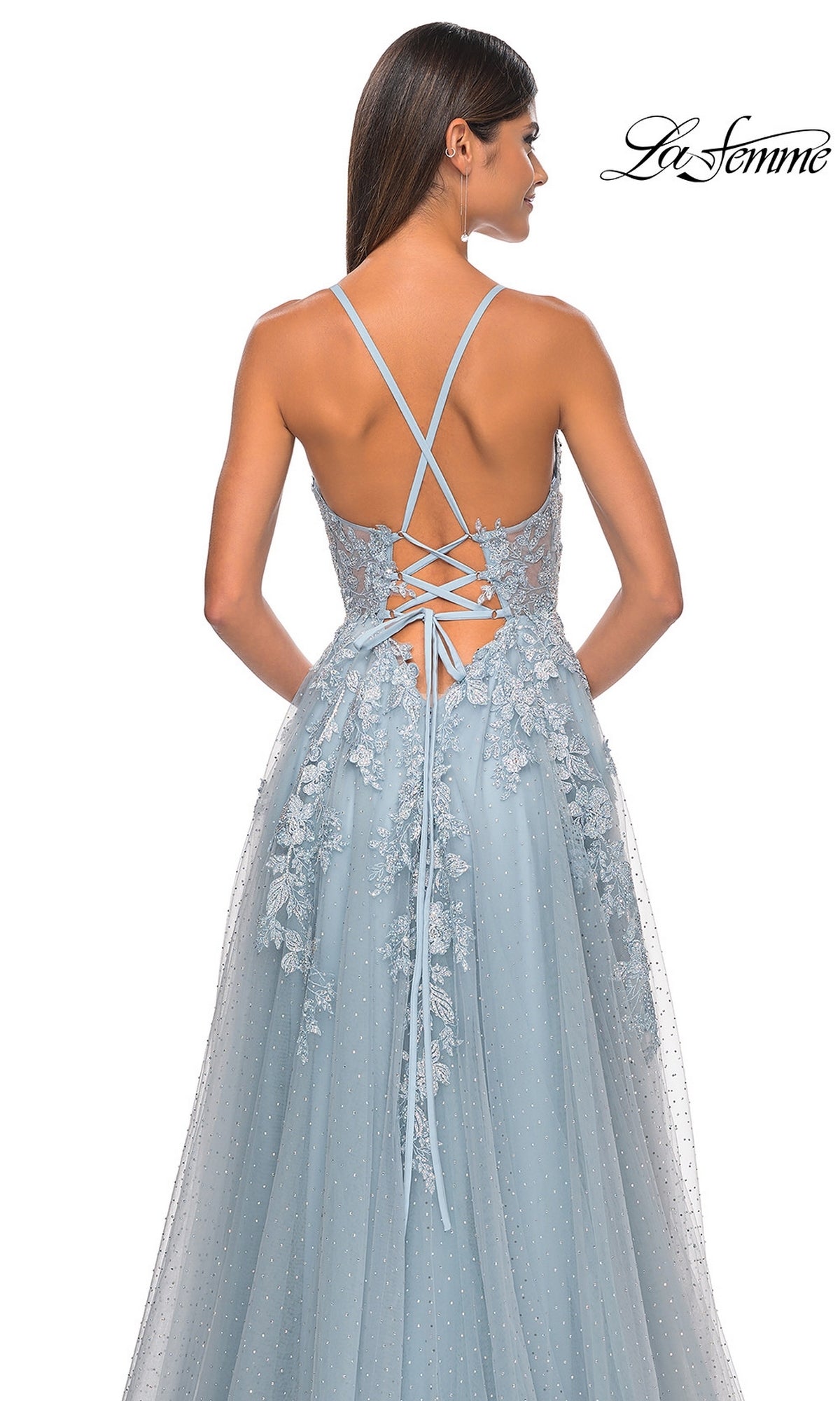 A woman wears the La Femme Lace-Embroidered Long Prom Dress 32147 in light blue with floral lace and tulle overlay, lace-up back, facing away from the camera. The